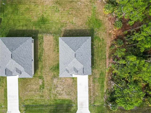 Aerial view featuring residential properties with hip roofs, concrete driveways, and expansive grassy lots bordered by dense mature trees
