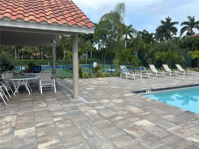 Poolside patio featuring multi-toned paver flooring, a covered seating area with a terracotta-style tiled roof, a swimming pool with a dark coping, and a tennis court visible in the background