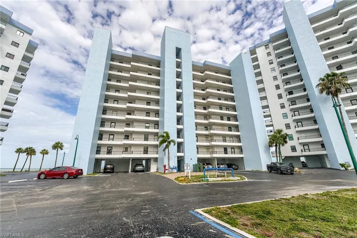 High-rise building with light blue and white exterior, multiple levels of balconies, and ground-level covered parking