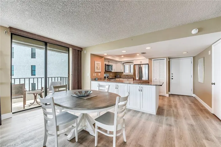 Open-concept living area with wood-finish flooring, a white cabinetry kitchen with light-colored countertops, a dining area featuring a round table, and a sliding glass door leading to a balcony with dark railings