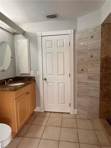 Bathroom featuring a wood-finish vanity with a stone countertop, a circular wall-mounted mirror, and a tiled shower enclosure