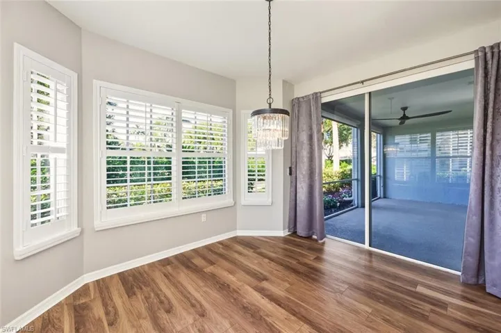 Wood-finish flooring throughout, featuring a crystal chandelier, multiple white plantation shutters, and sliding glass doors opening to a screened-in patio with a ceiling fan