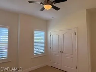 Neutral-toned interior featuring two windows with white plantation shutters, a double-door closet with arched paneling, and a ceiling fan with integrated lighting