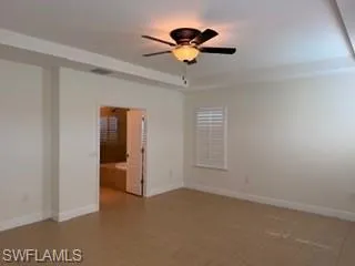 Spacious room featuring tile flooring, a ceiling fan with light fixture, tray ceiling, and a window with white plantation shutters