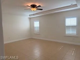 Spacious interior room featuring a tray ceiling with a ceiling fan light fixture, tile flooring, and two windows with plantation shutters