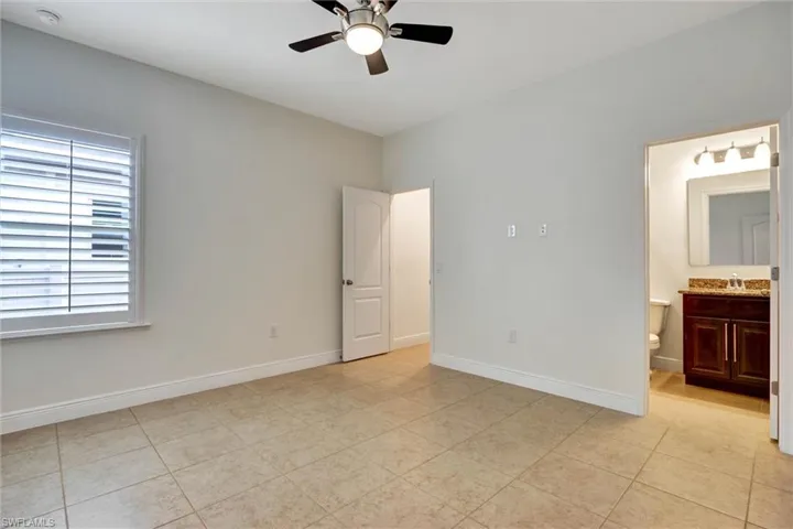 Neutral-toned room featuring tile flooring, a window with plantation shutters, a ceiling fan with integrated lighting, and a doorway opening to a bathroom with a vanity