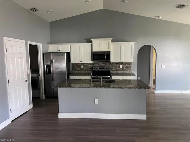 Kitchen featuring dark stone countertops, arched walkways, stainless steel appliances, white cabinets, and a center island with sink