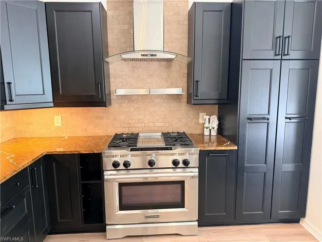 Kitchen featuring high end stainless steel range, wall chimney exhaust hood, tasteful backsplash, light stone countertops, and light wood finished floors