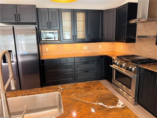 Kitchen featuring appliances with stainless steel finishes, glass insert cabinets, wall chimney range hood, and light stone countertops