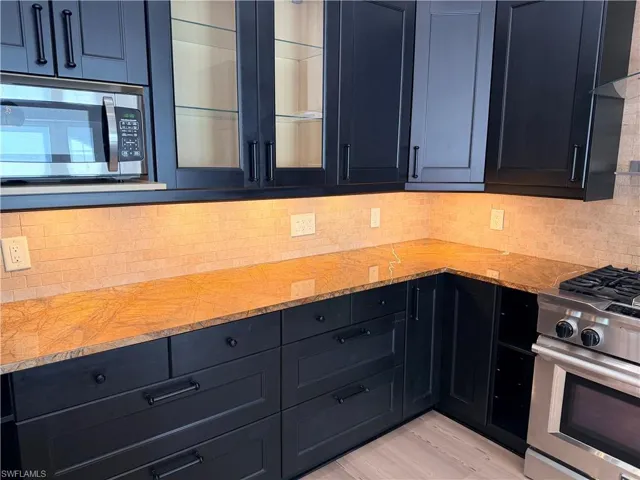 Kitchen with stainless steel appliances, glass insert cabinets, light stone countertops, dark cabinetry, and light wood-style flooring