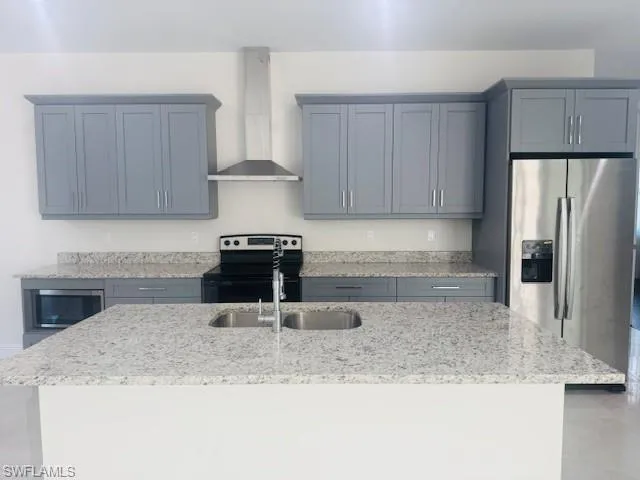 Kitchen featuring light stone countertops, appliances with stainless steel finishes, wall chimney exhaust hood, a kitchen island with sink, and gray cabinets