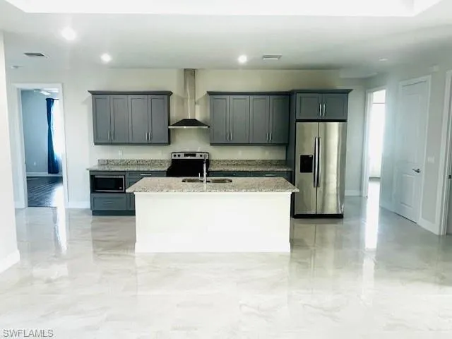 Kitchen with appliances with stainless steel finishes, wall chimney exhaust hood, an island with sink, light stone counters, and gray cabinetry
