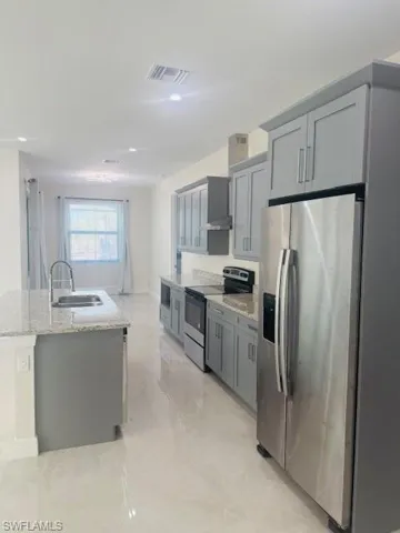 Kitchen featuring stainless steel appliances, light stone countertops, an island with sink, gray cabinets, and wall chimney range hood