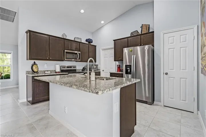 Kitchen featuring dark wood cabinetry, stainless steel appliances, vaulted ceiling, light stone counters, and an island with sink