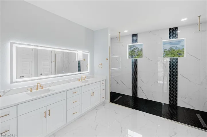 Spacious bathroom featuring a dual vanity with white cabinetry and gold-tone hardware, a large illuminated mirror, a walk-in shower with black hexagonal tile accents, and white veined tile flooring