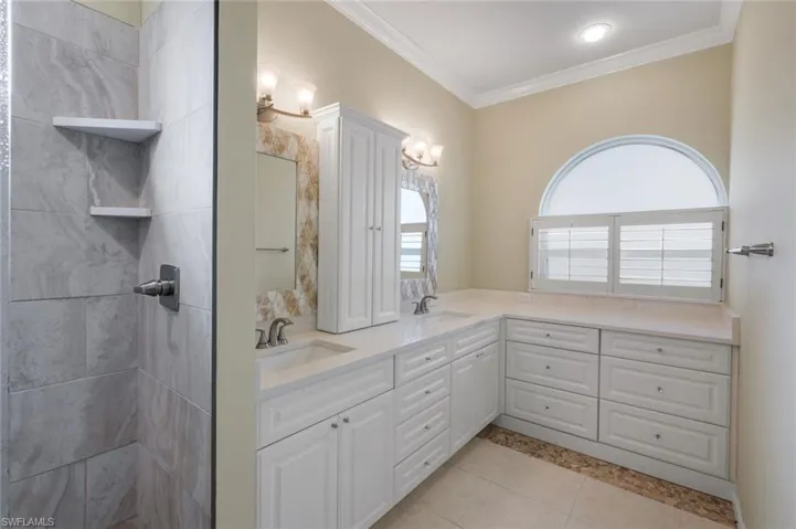 Dual vanity with white cabinetry and integrated sinks, a large arched window with shutter blinds, a tall built-in storage cabinet, and a tiled shower with integrated shelving