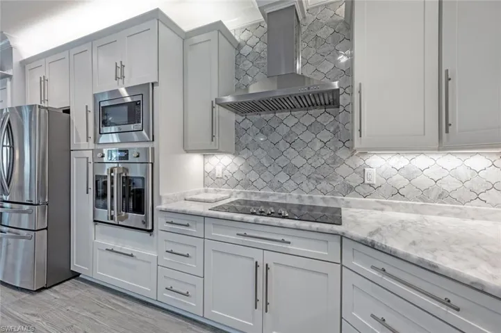 Kitchen featuring stainless steel appliances, wall chimney range hood, tasteful backsplash, light stone countertops, and light wood finished floors