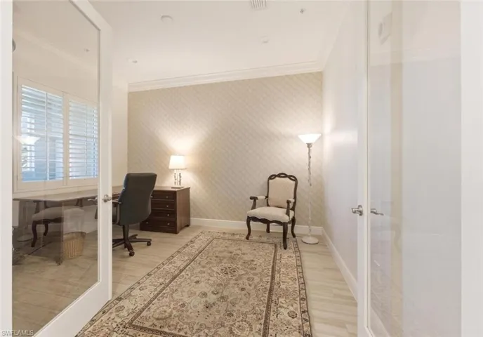Glass-paneled entry door opens to a room featuring light wood-finish flooring, patterned wallpaper accent wall, crown molding, and plantation shutters