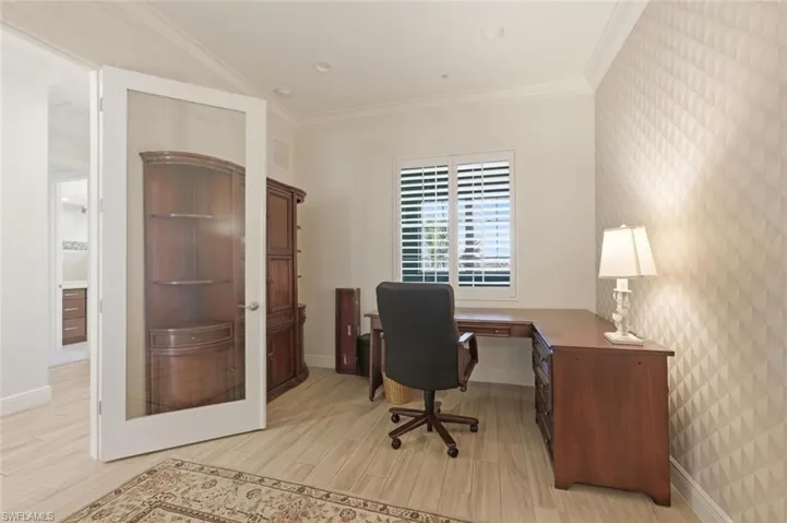Dedicated office space featuring wood-finish flooring, a full-lite glass door, crown molding, a window with plantation shutters, and a wall with geometric patterned wallpaper