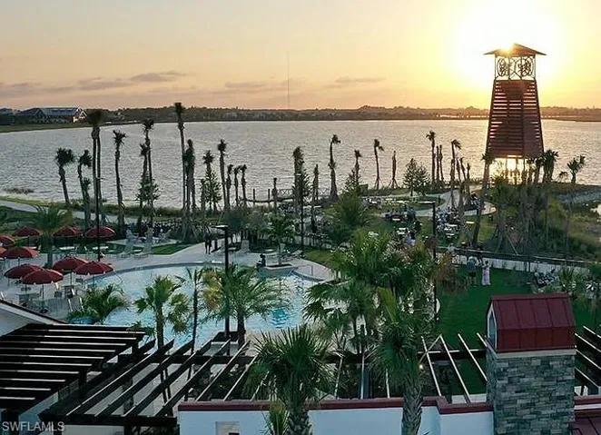 Expansive lakefront setting featuring a swimming pool with lounge seating, multiple palm trees, a decorative tower, and a stone-clad architectural element with a red roof