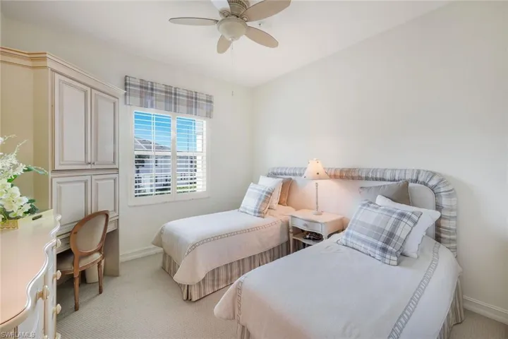 White-painted walls and light-colored carpeting define this bright room, featuring a window with white louvered shutters and a plaid valance