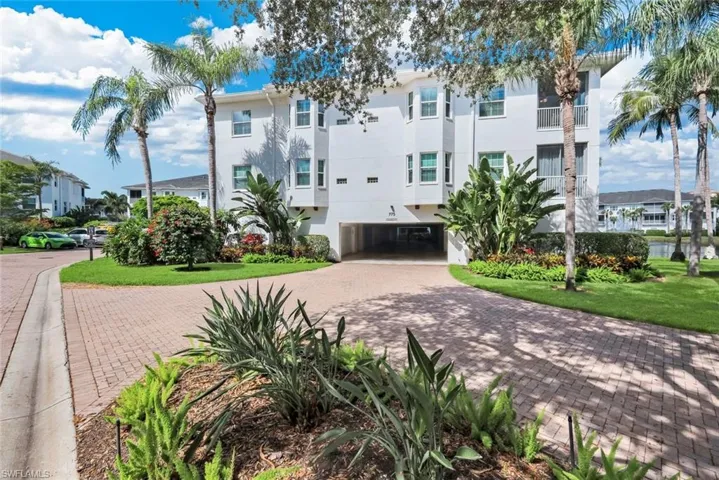 Paved brick driveway leading to a multi-story white stucco building with bay windows, mature palm trees, and lush landscaping