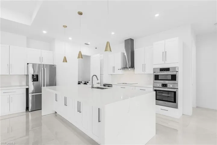 Bright kitchen featuring a large center island with integrated sink, white cabinetry, stainless steel appliances, and sleek pendant lighting
