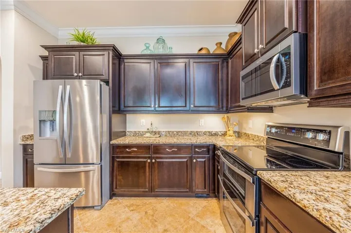 Kitchen with light tile patterned flooring, dark brown cabinets, stainless steel appliances, ornamental molding, and light stone countertops