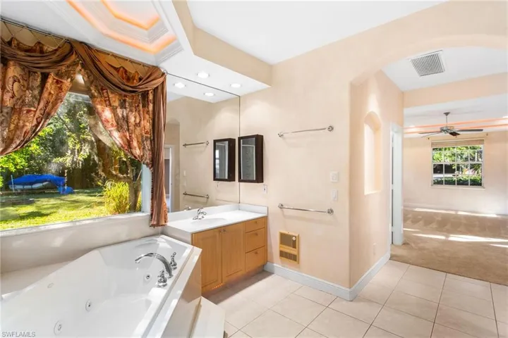 Bathroom featuring a built-in jetted tub, expansive window with outdoor scenery, double vanity with wood-finish cabinetry, full-wall mirror, and tray ceiling with recessed lighting