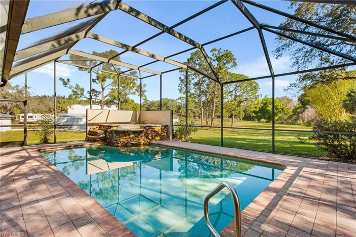 Screen-enclosed swimming pool featuring a stone-clad spa with tiered steps, brick paver decking, and a stainless steel grab rail