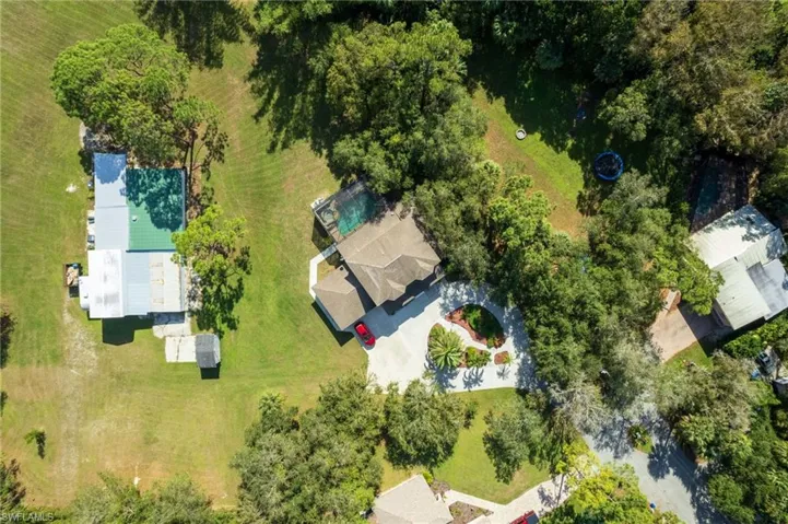 Aerial perspective showcasing a property featuring a brown shingle roof, a paved driveway, a manicured front lawn with mature landscaping, and an enclosed pool area