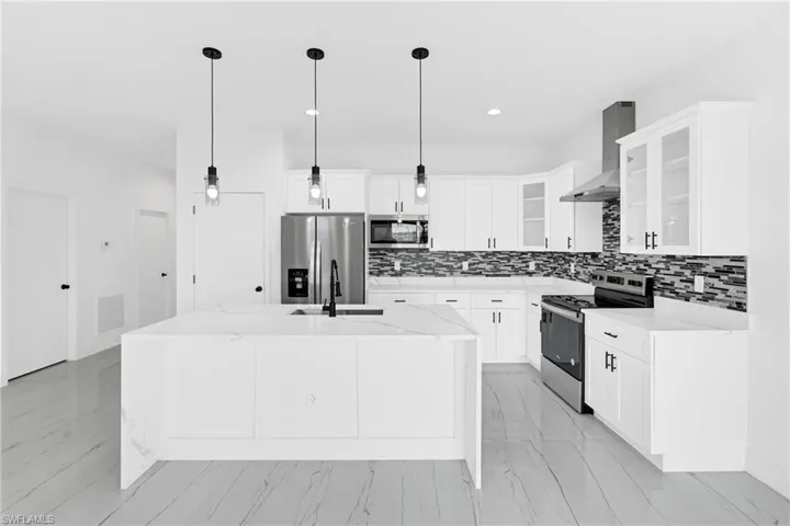 Modern kitchen featuring a waterfall-edge island with a built-in sink and pendant lighting, white cabinetry, a mosaic tile backsplash, and stainless steel appliances including a range hood