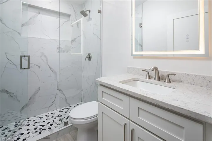The guest bathroom features a walk-in shower with white marbled wall tiles and stone tiled floor, sleek vanity with a light-colored quartz countertop, a rectangular sink and large mirror with illuminated border.