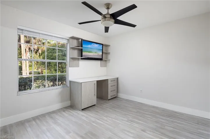 This bedroom features light-colored plank flooring, a window offering views of greenery, new baseboards, trim and interior door and a ceiling fan with a light fixture