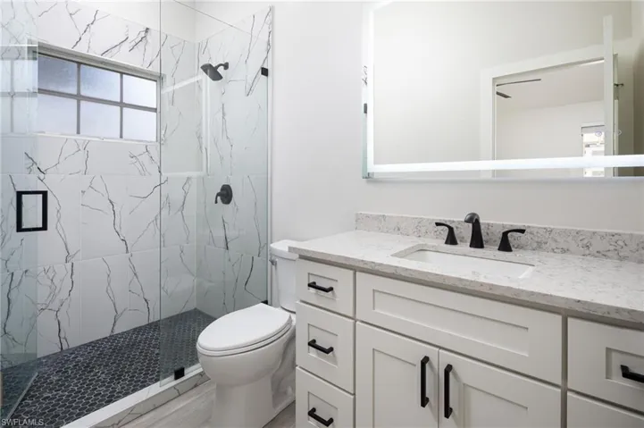 The primary bathroom featuring a walk-in shower with white marbled wall tiles and a dark hexagon tiled floor, a vanity with a white quartz countertop and sink, and a large mirror with an illuminated border