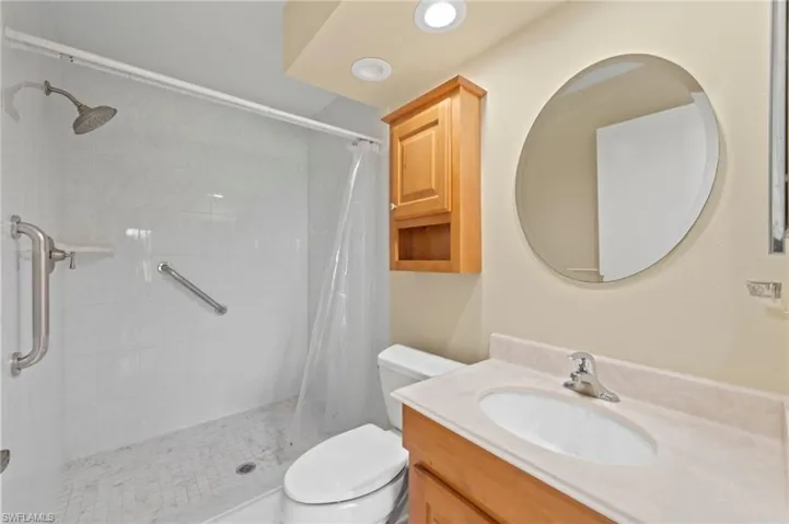 Bathroom featuring a walk-in shower with white tiled walls and grab bars, a round vanity mirror, a light wood-finish vanity with an integrated sink, and recessed ceiling lighting