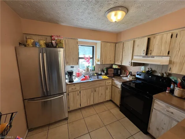 Kitchen with black electric range, freestanding refrigerator, light brown cabinets, light tile patterned floors, and under cabinet range hood