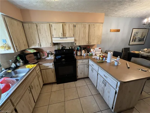 Kitchen with black appliances, a peninsula, a textured ceiling, light brown cabinets, and under cabinet range hood