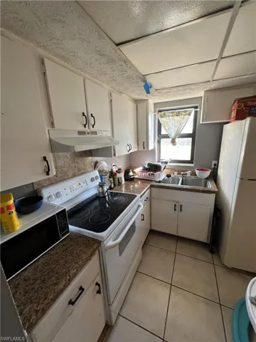 Kitchen featuring white appliances, white cabinetry, light tile patterned floors, under cabinet range hood, and dark countertops