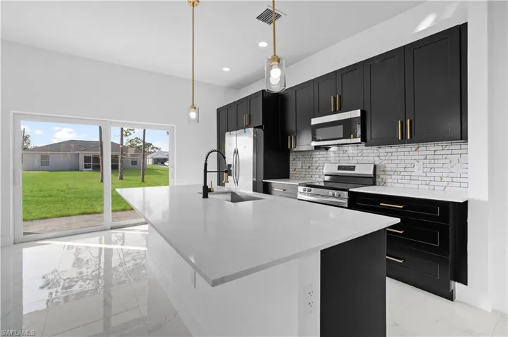 Kitchen with dark cabinetry, light marble finish flooring, stainless steel appliances, decorative backsplash, and an island with sink