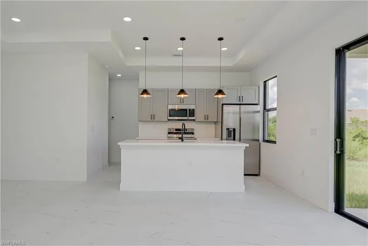 Kitchen with light marble finish flooring, appliances with stainless steel finishes, a raised ceiling, a center island with sink, and recessed lighting