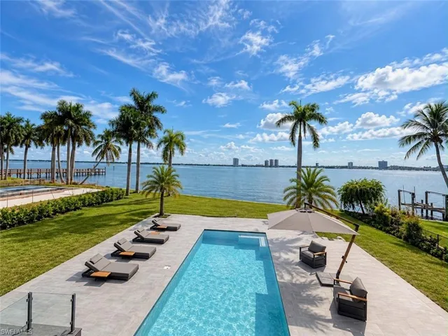 Expansive outdoor area featuring a swimming pool with a sun shelf, a surrounding stone tile patio, and a manicured lawn leading to a waterfront