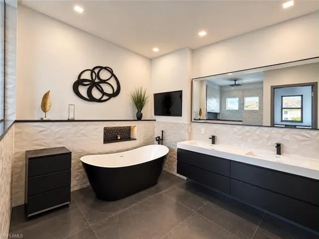 Freestanding soaking tub, dual floating vanity with integrated sinks, large wall-mounted mirror, recessed lighting, and dark tile flooring