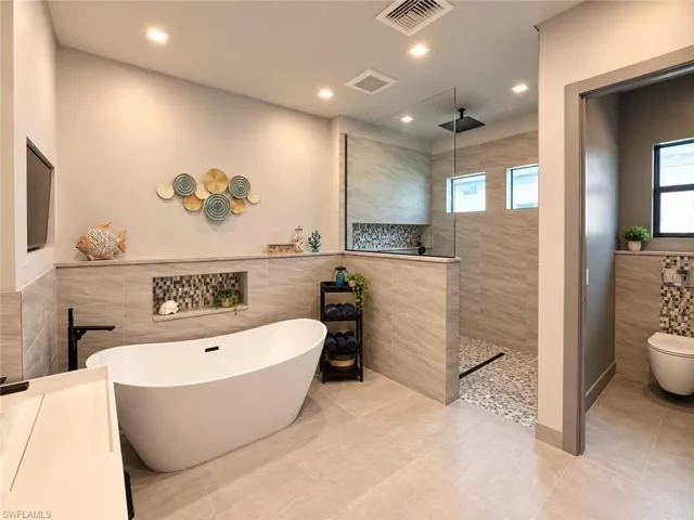 Freestanding soaking tub, walk-in shower with glass partition, tiled walls, large format tile flooring, and recessed lighting