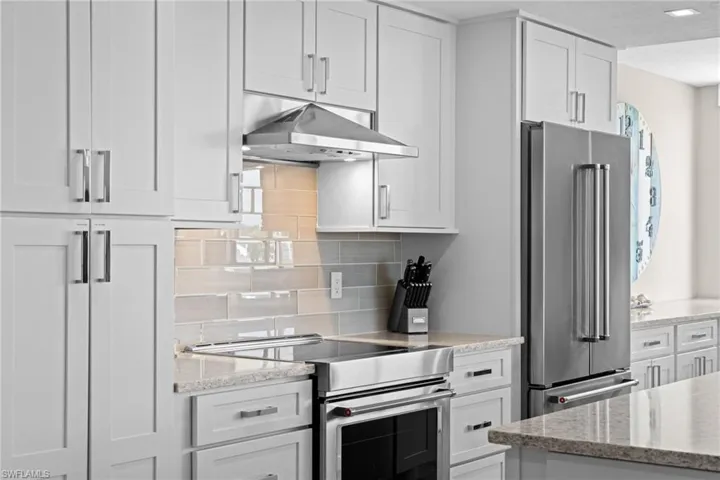 Modern kitchen featuring light gray shaker-style cabinetry, stainless steel appliances, a range hood, and a light-toned tile backsplash