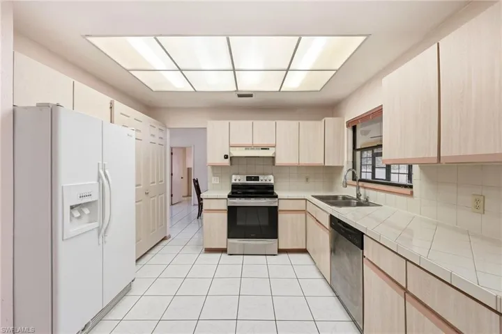 Kitchen featuring tile countertops, light brown cabinets, appliances with stainless steel finishes, decorative backsplash, and under cabinet range hood
