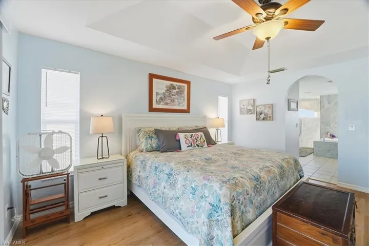 Bedroom with a tray ceiling, arched walkways, a ceiling fan, light wood-type flooring, and ensuite bath