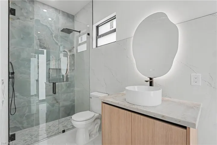 Modern bathroom featuring a frameless glass shower enclosure with rain shower head, light wood-finish vanity with countertop, a vessel sink, and an illuminated irregular-shaped mirror