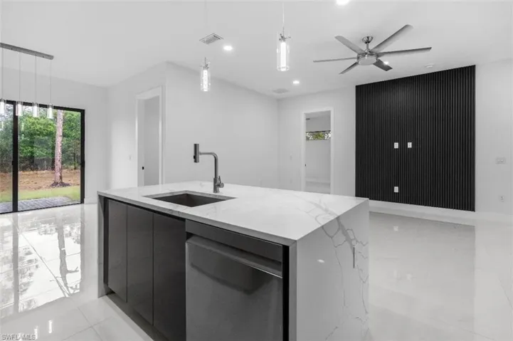 Kitchen with modern cabinets, light stone countertops, an island with sink, stainless steel dishwasher, and decorative light fixtures
