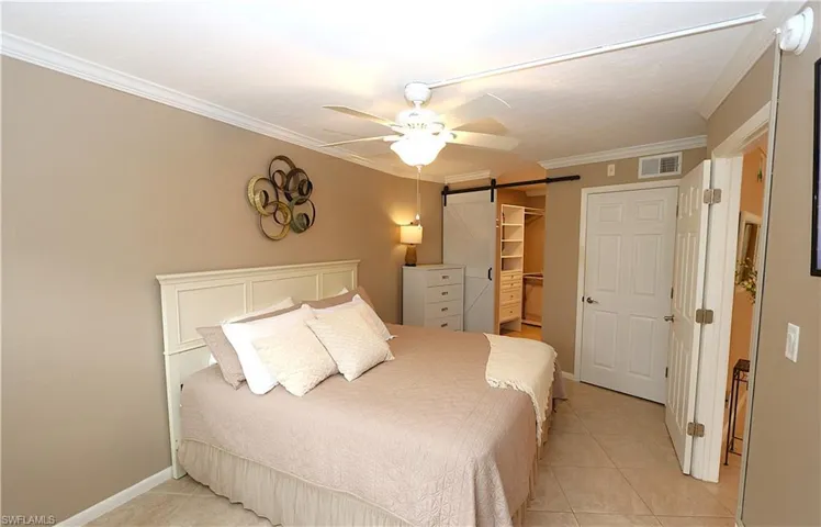 Bedroom with a barn door, connected bathroom, crown molding, light tile patterned floors, and ceiling fan